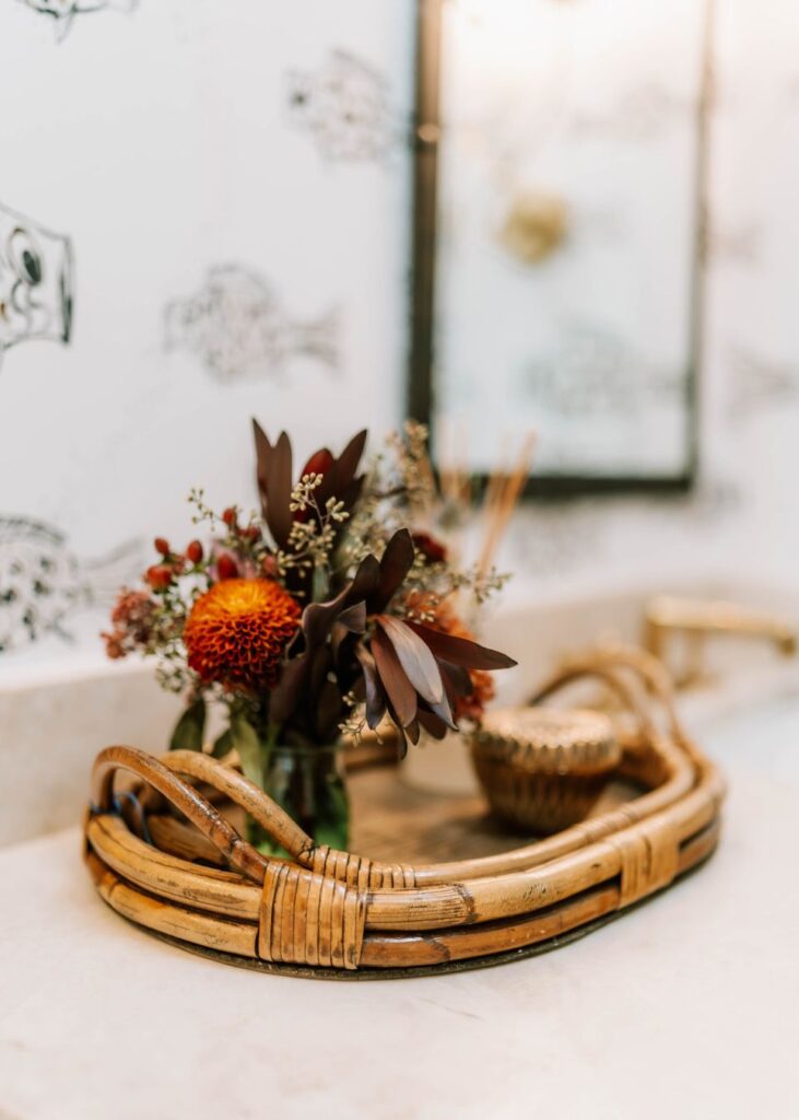 A rattan tray on a bathroom counter holds a small vase with dark red and orange flowers and a decorative container. Styled by an interior decorator in Boerne, the background features a black-and-white fish pattern and a blurred mirror.