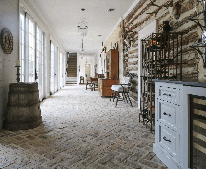 A rustic hallway with exposed log walls, brick flooring, large windows on the left, a wooden barrel, antique furniture, wine racks, and mounted antlers, leading to a staircase at the far end.