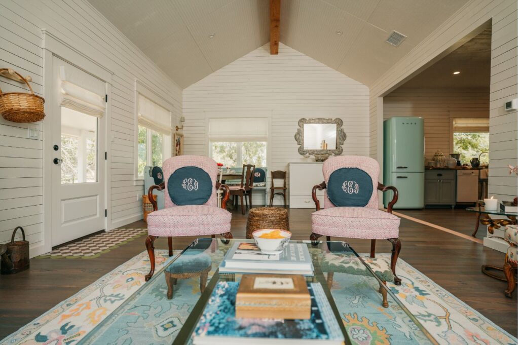 Cozy living room styled by an interior designer Boerne TX, featuring two pink armchairs, monogrammed pillows, a decorative rug, coffee table with books, and a kitchen view with retro mint fridge. Light pours through large windows.
