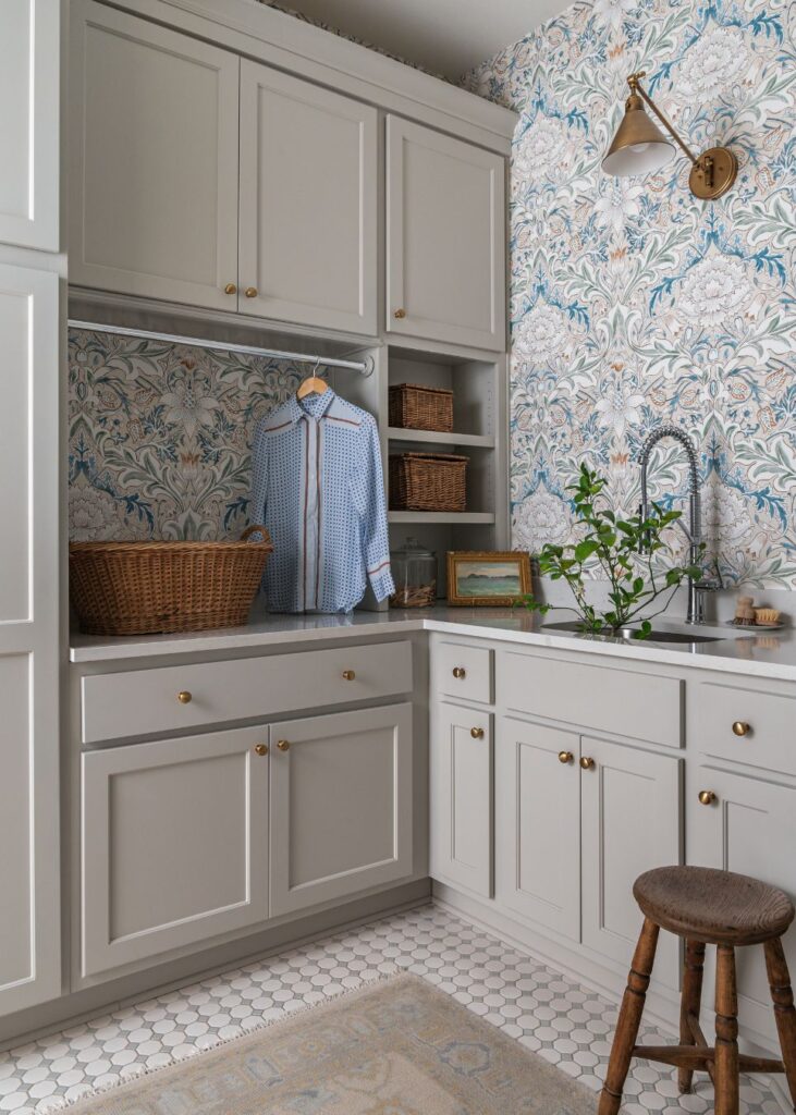 A laundry room with white cabinets, patterned wallpaper, a wicker basket, a blue shirt hanging, open shelves with baskets—a charming space full of laundry room design ideas inspired by modern farmhouse interior design.