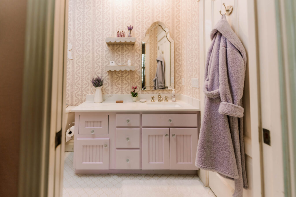 A bathroom with pink cabinets, a decorative mirror, potted plants, and shelves with toiletries styled by an interior designer Boerne TX. A lavender bathrobe hangs on a wall hook; vintage wallpaper and white tile floors complete the look.