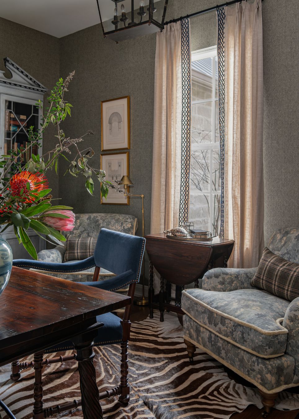 A cozy sitting room, curated by an interior decorator in Boerne, features two patterned armchairs with plaid cushions, a dark wood table, animal print rug, framed art, and tall curtains framing a window aglow with soft light.