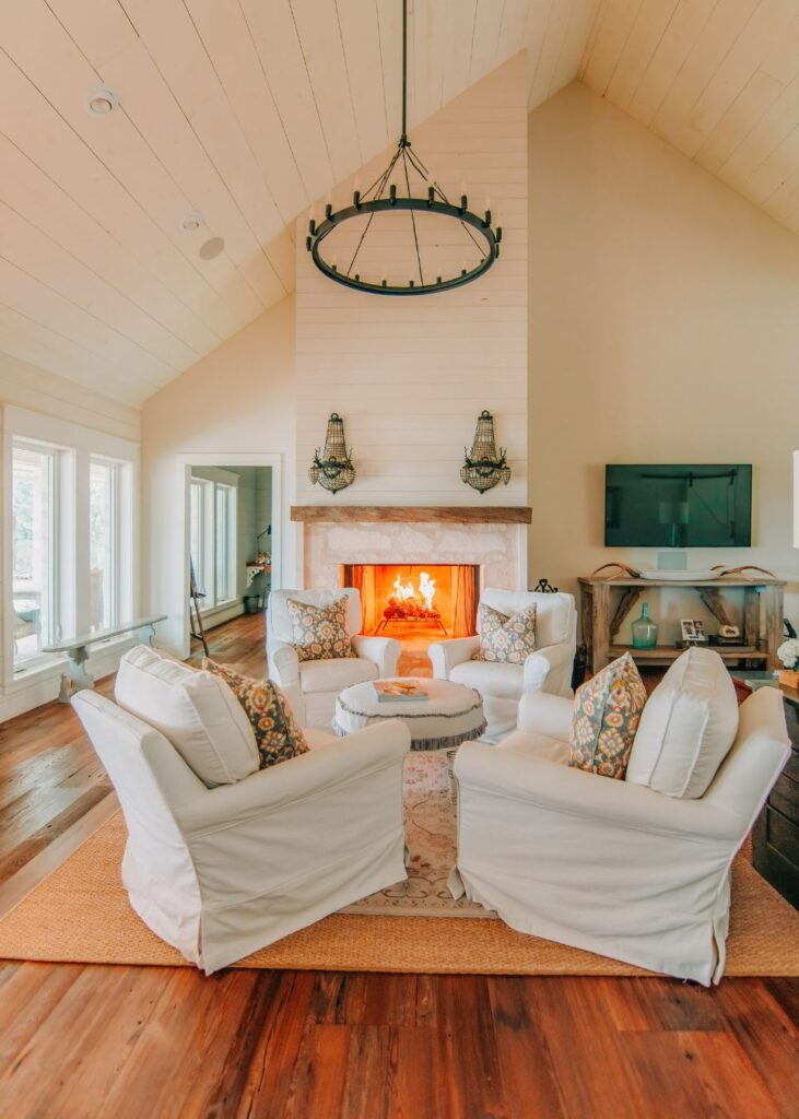 Bright, cozy living room with vaulted ceiling, styled by an interior design company Boerne TX; four white armchairs around a small table, a stone fireplace with a lit fire, wall sconces, TV, and large windows letting in natural light.