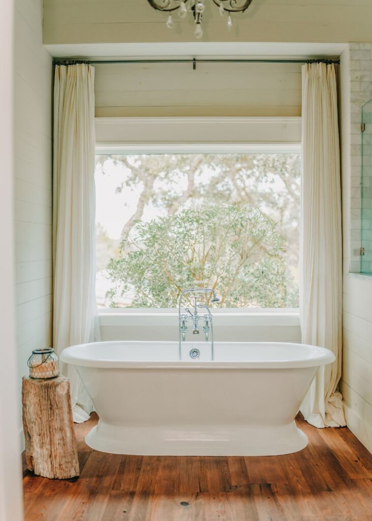 A white freestanding bathtub is centered in front of a large window with cream curtains, overlooking greenery—an inviting retreat crafted by an expert interior designer Boerne TX, with wooden floors and a rustic wooden stool adding warmth.