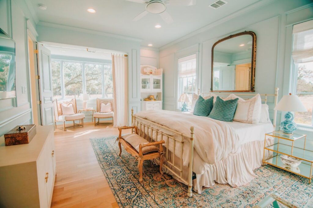 A bright, elegant bedroom with pale blue walls, a large bed with white and blue pillows, a decorative rug, and a gold-framed mirror—crafted by an expert interior designer Boerne TX. Sunlit windows and a cozy sitting area complete the space.