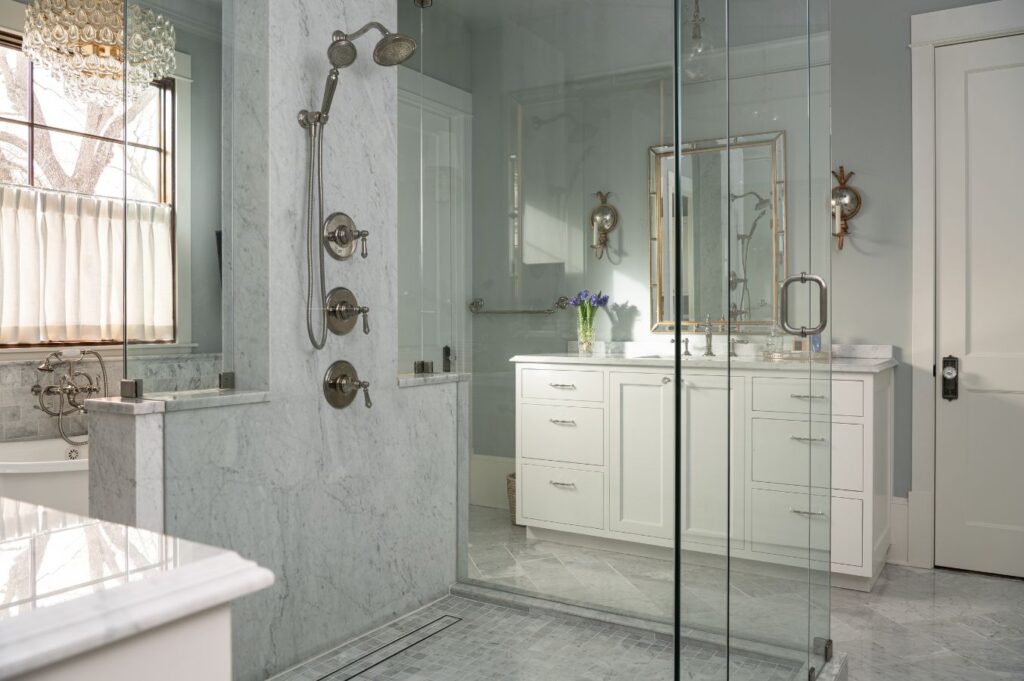 Bright, elegant bathroom designed by a top interior designer Boerne TX, featuring a glass walk-in shower, marble walls and floor, white vanity with mirror, chrome fixtures, window with sheer curtains, and fresh flowers on the vanity.