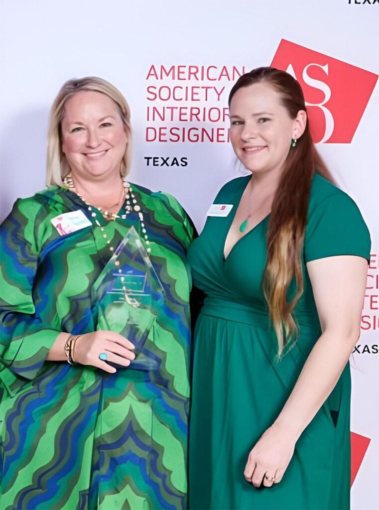Two women in green dresses smile at the camera; one holds a glass award. They stand in front of an American Society of Interior Designers Texas backdrop, representing an acclaimed interior design company Boerne TX.