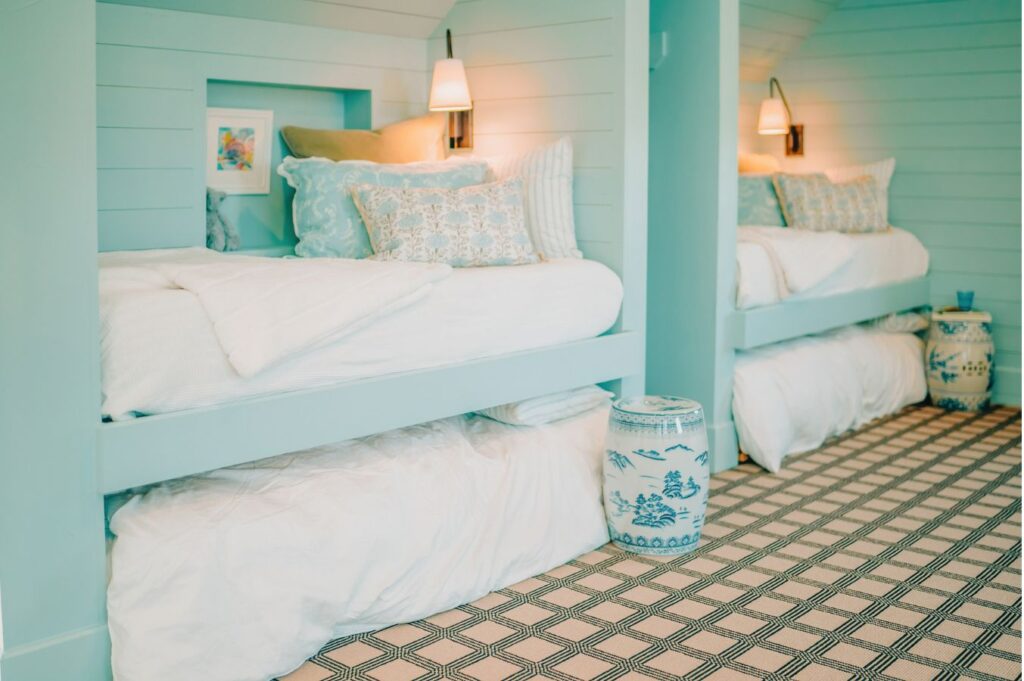 Two light blue built-in bunk beds with white bedding and decorative pillows create a cozy atmosphere. Soft wall lights, a blue-patterned ceramic stool, and geometric-patterned carpet showcase the work of an expert interior designer Boerne TX.