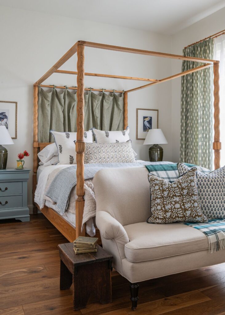 A cozy bedroom styled by an interior decorator in Boerne, featuring a wooden four-poster bed, layered pillows and blankets, a beige loveseat at the foot, blue nightstands with lamps, and patterned curtains. Sunlight streams through the window.