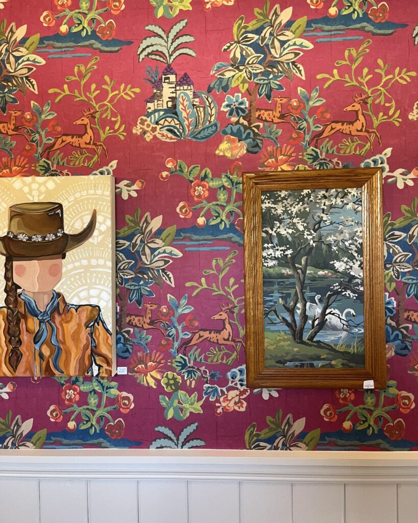 Two framed paintings hang on vibrant floral wallpaper, styled by an interior designer Boerne TX. The left painting shows a braided figure in a cowboy hat; the right displays a blooming tree by water. White wainscoting completes the look.
