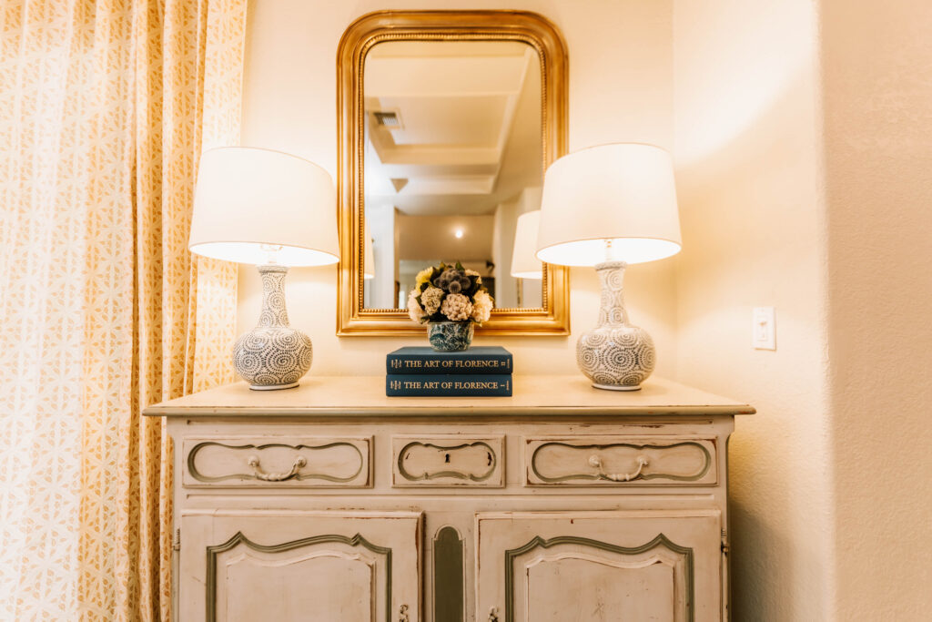 A vintage cream-colored dresser with ornate details holds two white table lamps, a stack of books titled The Art of Florence, and a small bouquet. Styled by an interior designer Boerne TX, a gold-framed mirror hangs above, reflecting the room behind.