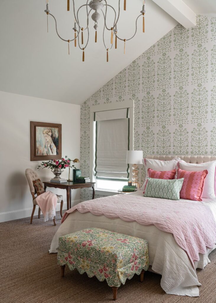 A cozy bedroom with a pink and green color scheme, expertly styled by an interior designer Boerne TX, features pink pillows, a scalloped quilt, floral bench, patterned accent wall, chandelier, and a small desk adorned with flowers and art.