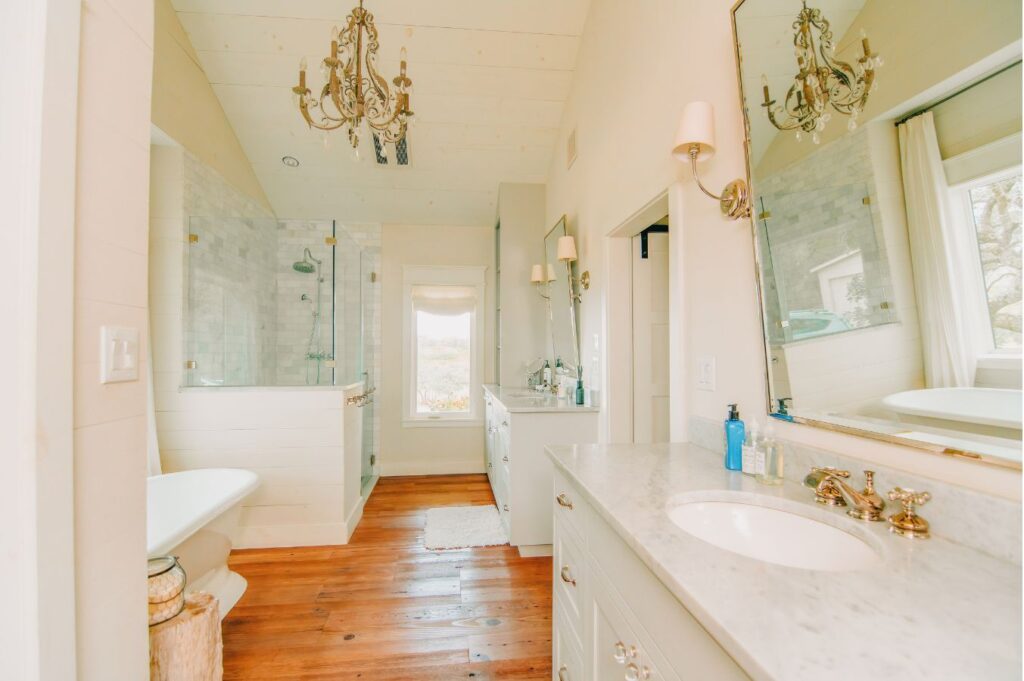 Bright, elegant bathroom with a marble countertop, double sink, freestanding tub, large mirrors, chandeliers, wooden floor, and a glass-enclosed shower—expertly curated by an interior design company Boerne TX. Natural light streams in from a window at the far end.