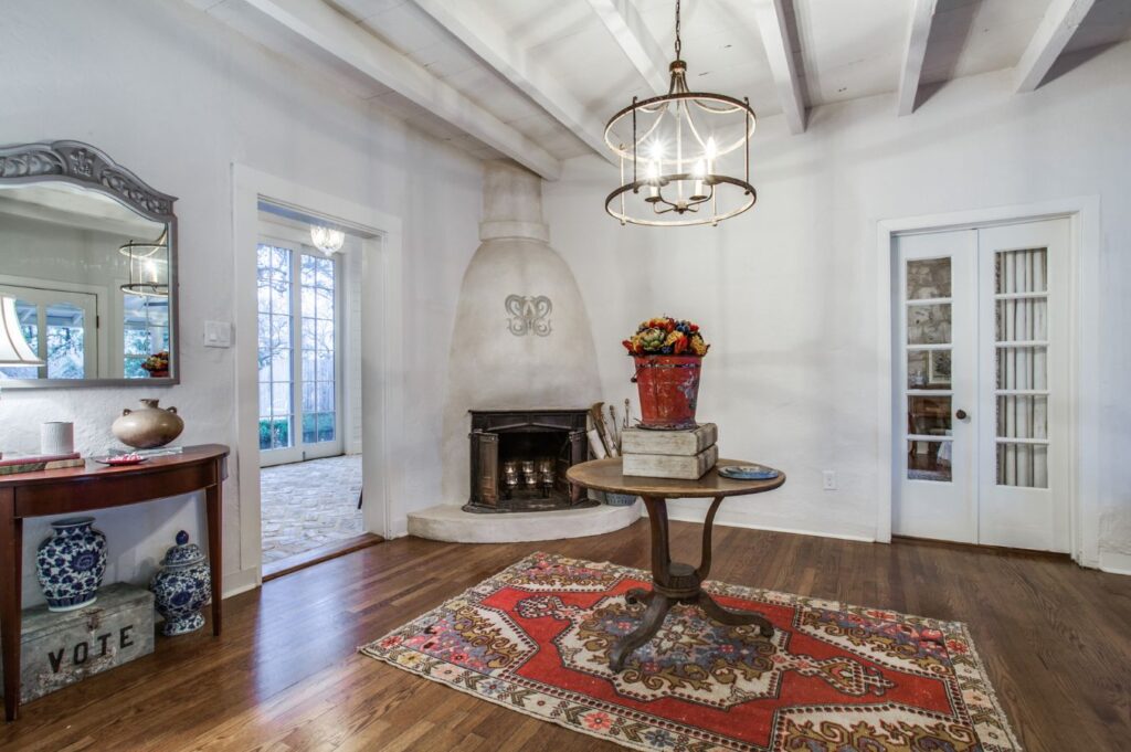 A cozy room with white walls, wood floor, a round table on a red patterned rug, a decorative fireplace, chandelier, side table with blue vases, and glass doors letting in natural light—perfect inspiration from an interior design company Boerne TX.