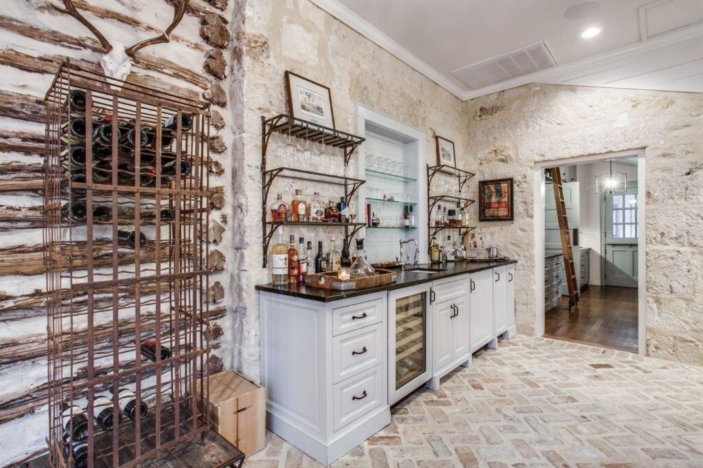 A rustic home bar area with stone walls, a wine rack filled with bottles, white cabinets, a small sink, open shelves for liquor and glasses, and a brick-patterned floor—expertly curated by an interior designer Boerne TX. A hallway leads to another room.