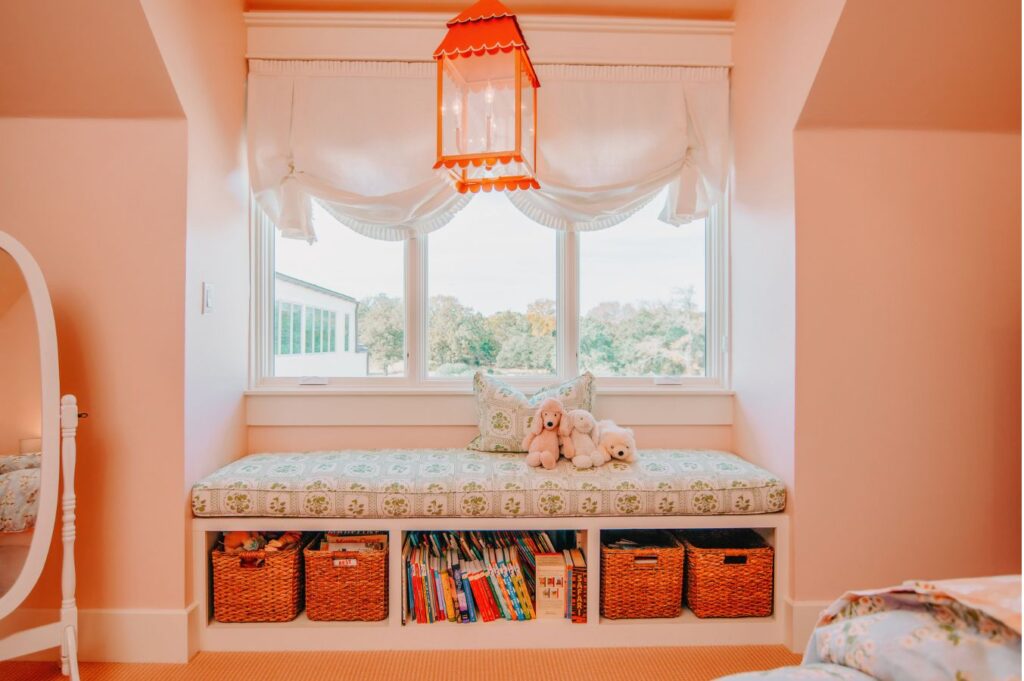A cozy window seat with patterned cushions, stuffed animals, and pillows. Below are shelves with baskets and children's books—an inviting nook designed by an interior designer Boerne TX. Bright natural light pours in through large windows and an orange lantern glows above.