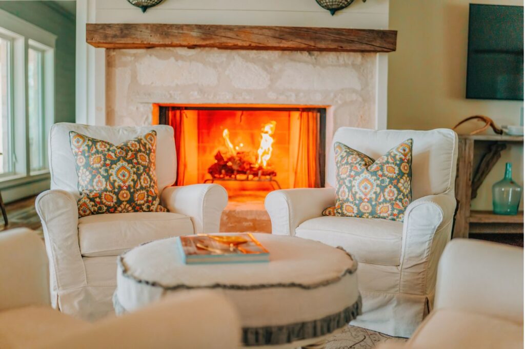 Two white armchairs with patterned pillows face a lit stone fireplace. A round ottoman with a book sits between them—an inviting living room crafted by an expert interior designer Boerne TX, filled with warm, natural light from nearby windows.