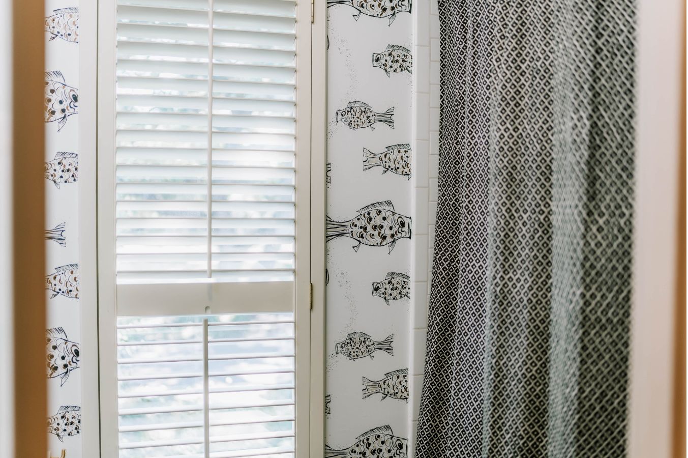 A window with white shutters is partially open. The wall is adorned with fish-themed wallpaper, and a bold black-and-white curtain hangs to the right—a charming touch from an interior designer in Boerne, TX.