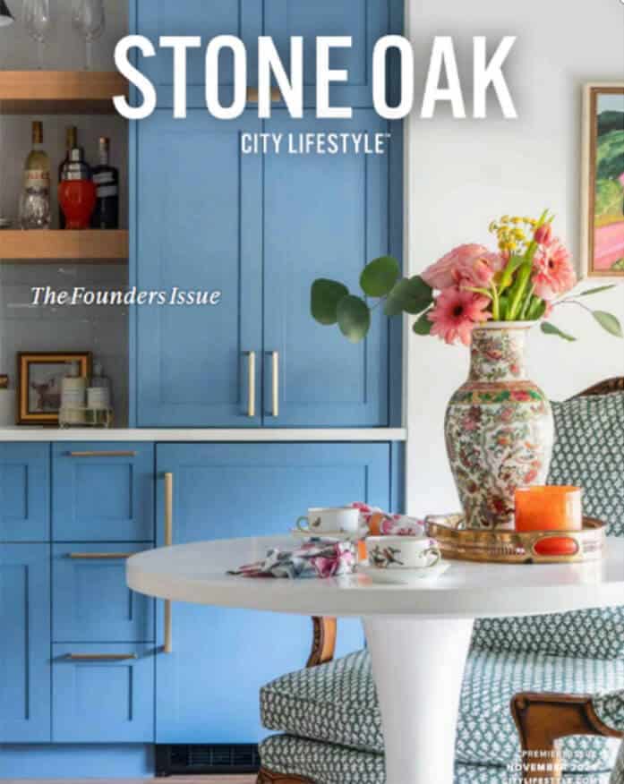 A magazine cover featuring blue kitchen cabinets, a white table set with floral teacups, a vase of pink flowers, and an upholstered chair—styled by an interior design company Boerne TX. The title reads STONE OAK City Lifestyle®—The Founders Issue.