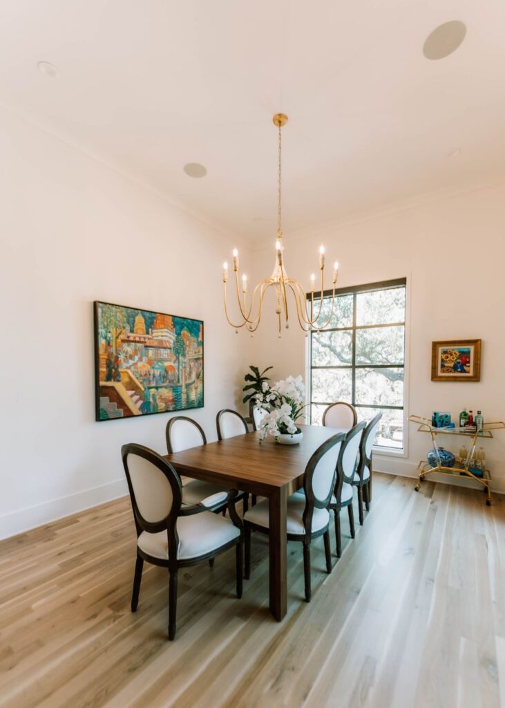 A bright dining room designed by an interior designer Boerne TX features a wooden table, six upholstered chairs, a gold chandelier, art on the walls, natural light from a window, a plant, and a bar cart with bottles and glasses.