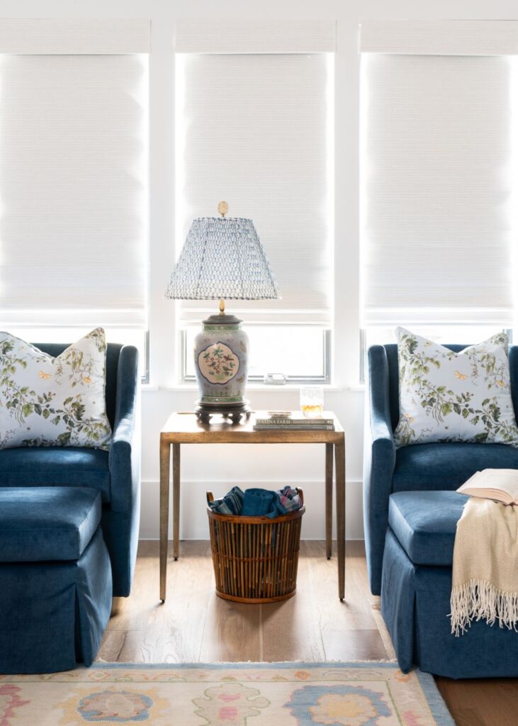 Two blue armchairs with floral pillows sit beside a small table topped with a lamp and glass of water. A basket of books rests below. White blinds cover bright windows, showcasing the cozy style from an interior design company in Boerne, TX.