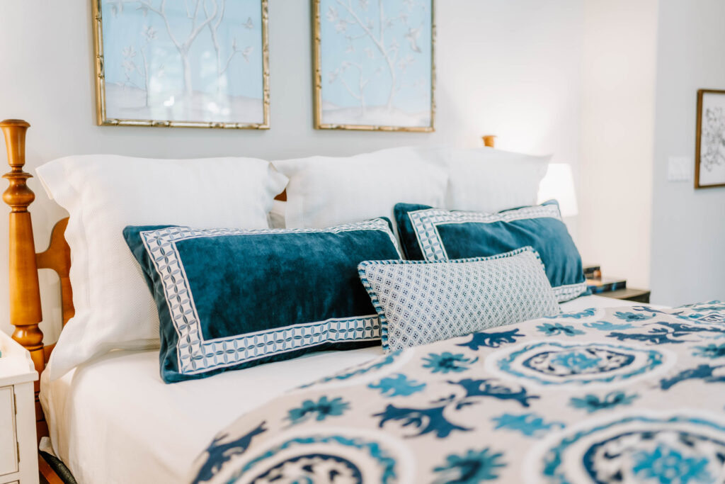 A neatly made bed with white and blue accent pillows and a matching patterned blanket showcases the touch of an interior decorator Boerne. Two framed artworks hang above the headboard, with a nightstand and lamp to the side.
