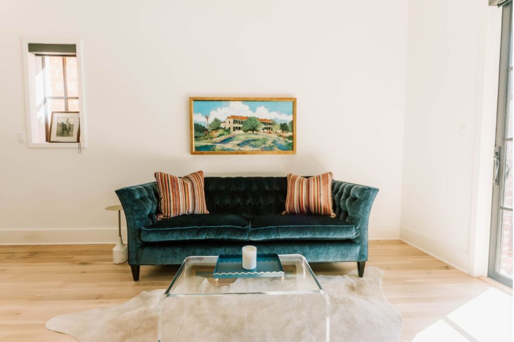 A modern living room by an interior designer in Boerne, TX, featuring a dark teal velvet sofa with striped pillows, a glass coffee table, white rug, wall art above the sofa, light wood floors, a small window, and a framed photo on the left wall.