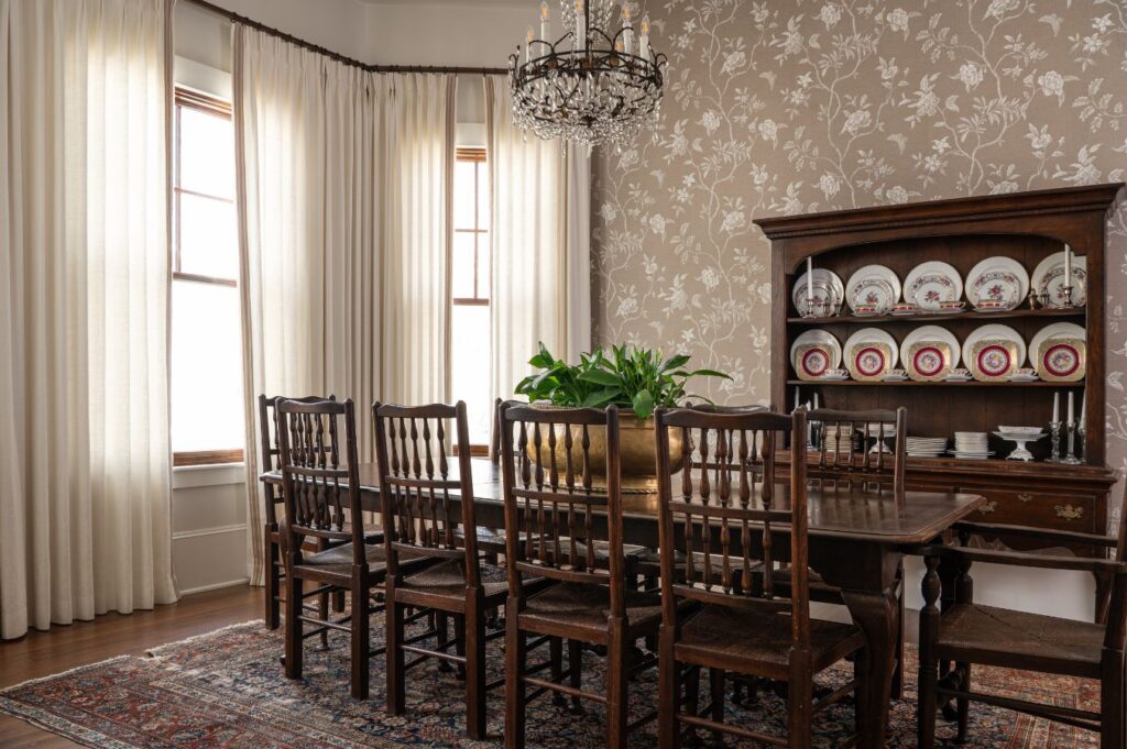 A traditional dining room by an interior decorator Boerne features a long wooden table, eight chairs, a chandelier, floral wallpaper, cream curtains, a rug, and a dark hutch with decorative plates. A potted plant adds charm to the classic setting.