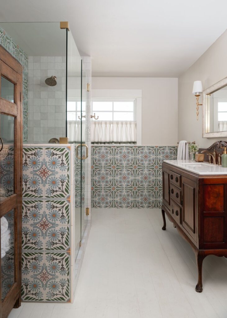 Vintage-style bathroom with patterned tiles, a glass shower enclosure, wooden vanity with marble countertop, wall sconce, and a window with sheer white curtains—perfect inspiration from an interior designer Boerne TX trusts for creating bright, welcoming spaces.