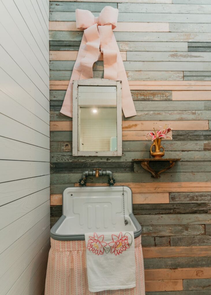 A rustic bathroom with a white sink, pink patterned skirt, embroidered towel, and wood-paneled wall is beautifully styled by an interior design company in Boerne, TX—complete with a pink bow and shelf holding fresh flowers.