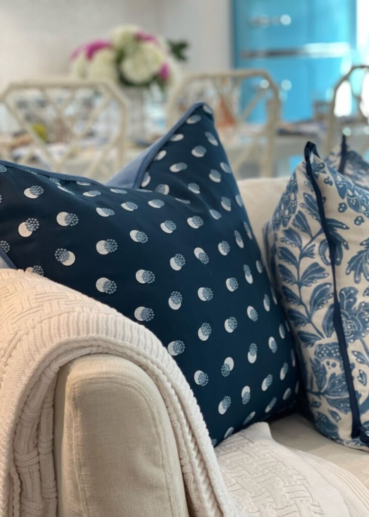 A beige sofa draped with a textured cream blanket features two decorative blue pillows—one with white polka dots and another with a blue floral pattern, perfectly styled by an interior design company in Boerne, TX. A blurred dining area is visible in the background.