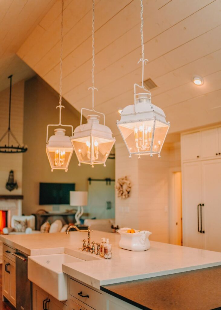 A modern farmhouse kitchen styled by an expert interior designer Boerne TX, featuring three white lantern-style pendant lights over a marble island with a farmhouse sink, stainless steel appliances, and warm, cozy lighting.