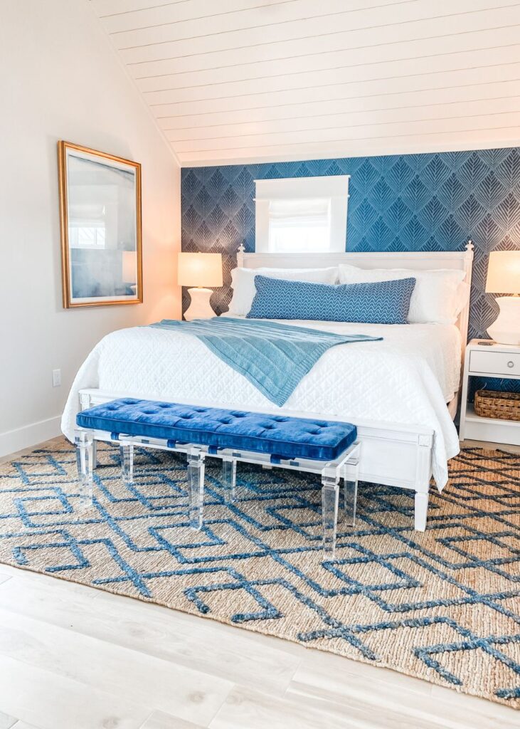 A bright bedroom with a white bed, blue and white bedding, and a textured blue accent wall—styled by an interior designer Boerne TX. The space features a geometric blue rug, clear-legged bench, and two softly lit bedside lamps.