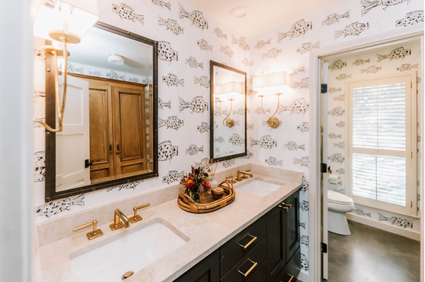 Bright bathroom with double sinks, gold fixtures, two mirrors, and fish-patterned wallpaper. Designed by an interior designer Boerne TX, a tray with flowers adds charm. A toilet and window with blinds are visible in the adjacent room.