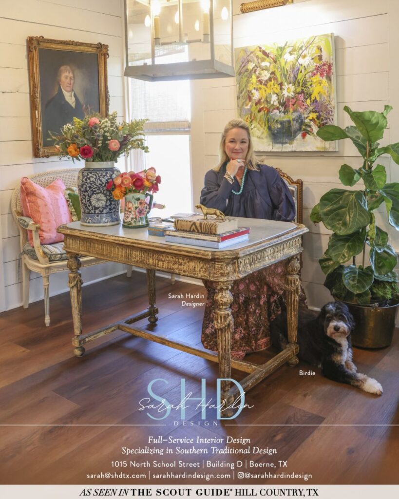 A woman sits at an ornate desk in a stylish office designed by an interior decorator Boerne, with a gold-framed portrait, plants, and a black dog on a patterned rug. The chic space features white shiplap walls and wood floors.