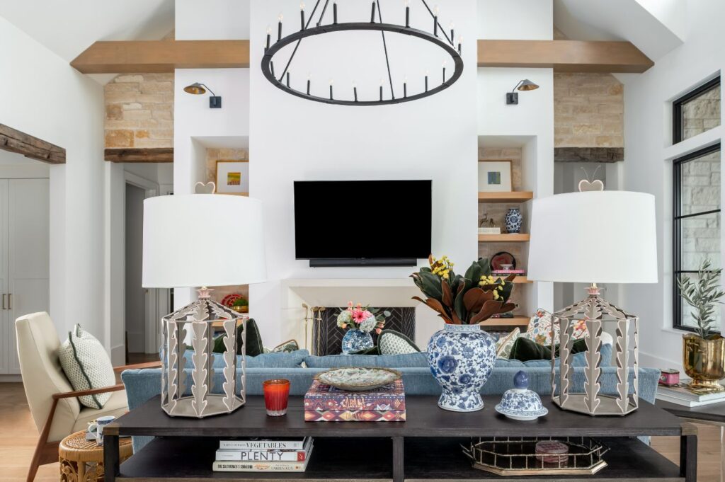 A modern living room by an interior design company in Boerne, TX, featuring a blue sofa, round chandelier, decorative lamps, vases, books on a black console table, wooden shelves, and windows flanking a TV on a white wall.
