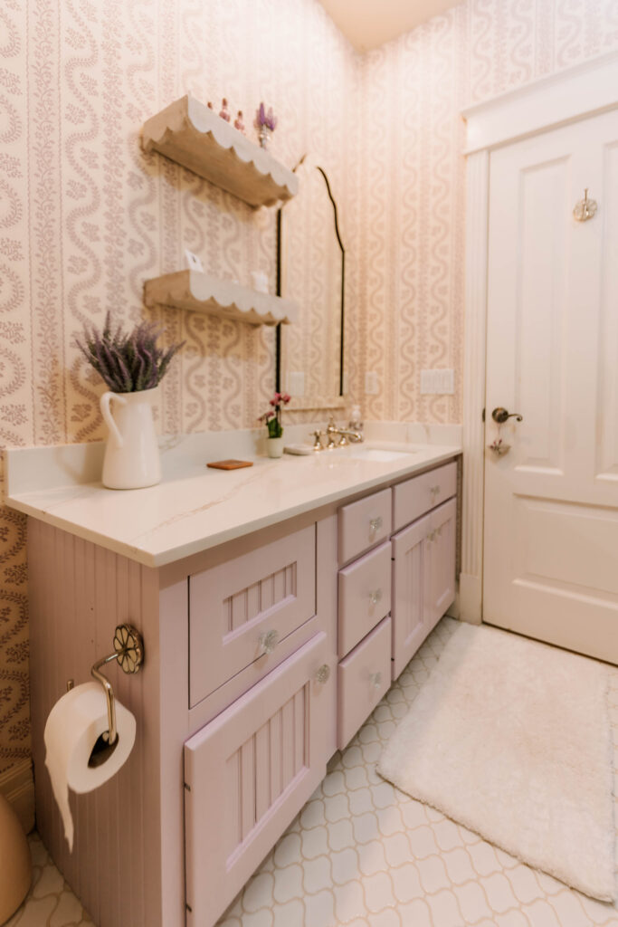 A bathroom by an interior designer Boerne TX features pale pink cabinets, a white countertop, a scalloped mirror, and open shelves with decor. A white pitcher with lavender sits on the counter against light, intricately patterned wallpaper.