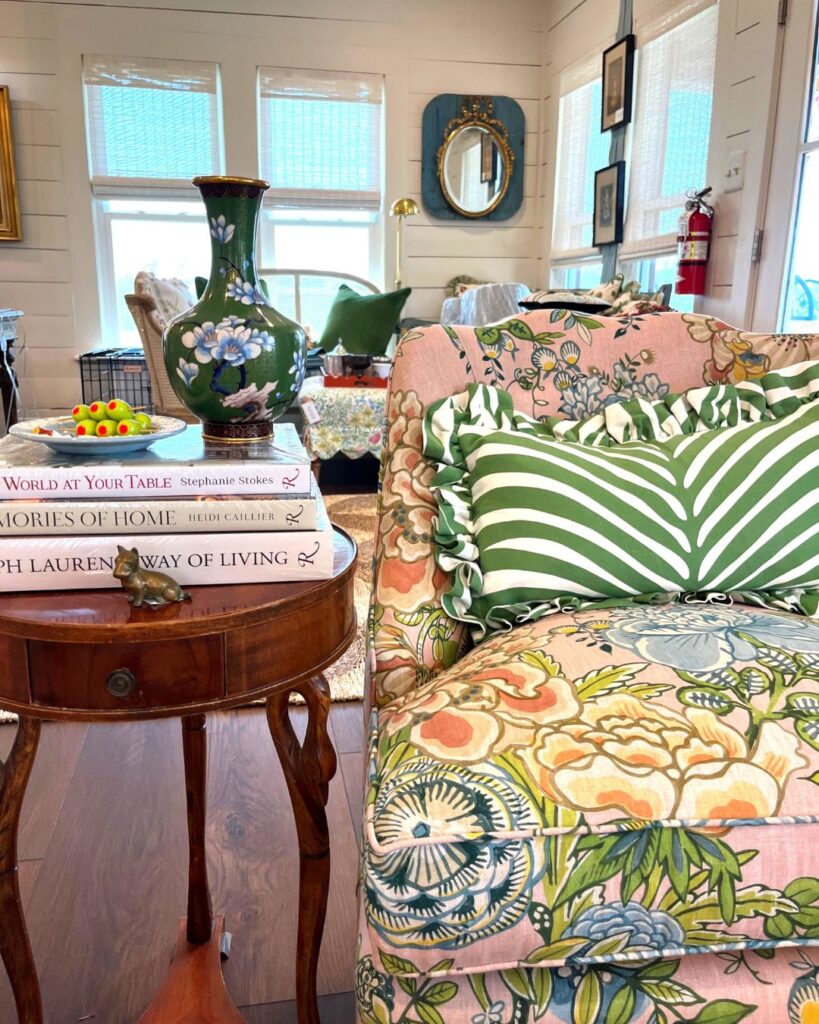 A cozy living room scene, curated by an interior decorator Boerne, with a floral armchair, green-striped pillow, and round wooden table holding a vase and books. Windows, mirrors, and eclectic decor complete this inviting space.