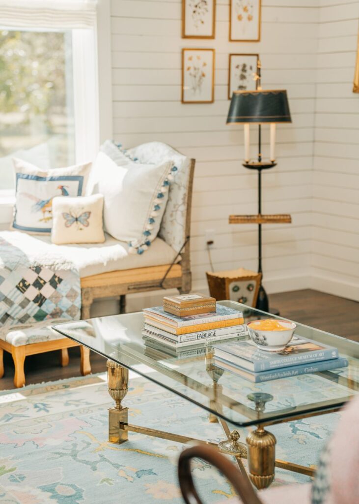 A sunlit living room styled by an interior designer Boerne TX features a glass coffee table with stacked books and a bowl, a cushioned bench with quilts and decorative pillows, a standing lamp, and framed art on white paneled walls.