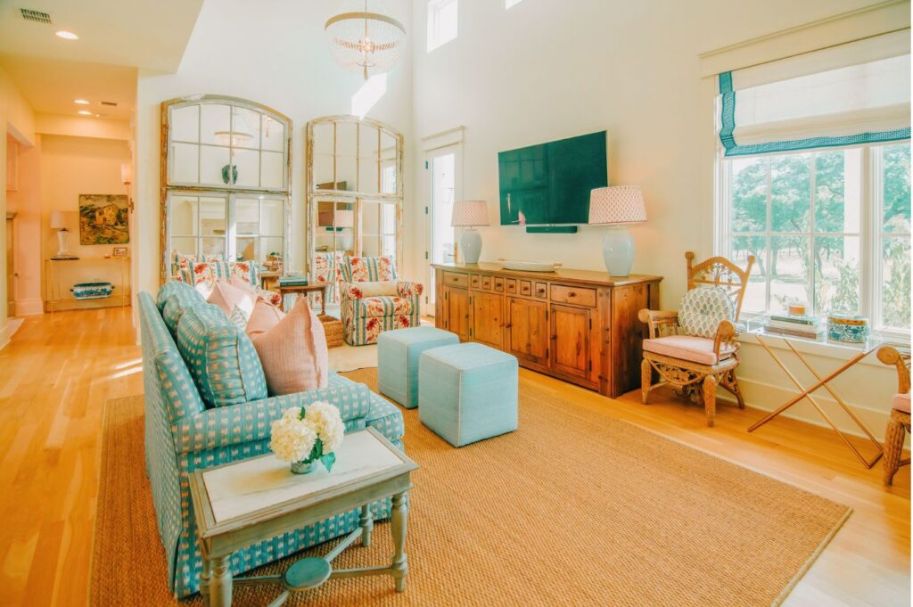 Bright living room with light wood floors, a blue sofa, patterned armchairs, two ottomans, a wooden console and TV—styled by an expert interior designer in Boerne TX. Large windows, woven rug, and mirrors complete the look.