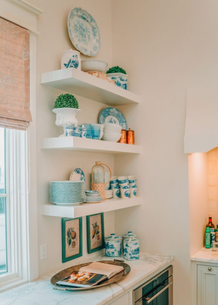 A kitchen corner styled by an interior design company in Boerne, TX, features white floating shelves with blue and white china, stacked plates, cups, small potted plants, marble counters below, an open magazine tray, and framed wall art.