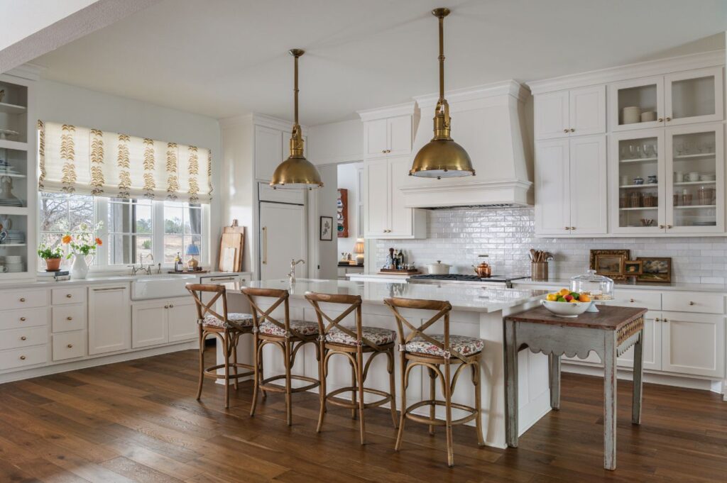A bright, spacious kitchen by a historic home interior designer features white cabinets, a large island with four wooden stools, brass pendant lights, wooden floors, and a farmhouse table. Sunlight streams through windows with patterned shades. Fruit and plants adorn the space.