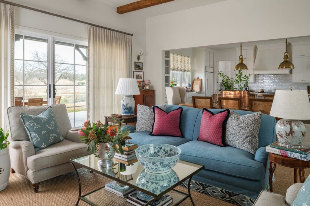Cozy living room with a blue sofa, red and patterned pillows, a glass coffee table with books and a decorative bowl, beige armchair, and view into a kitchen and dining area with wooden furniture and pendant lights.