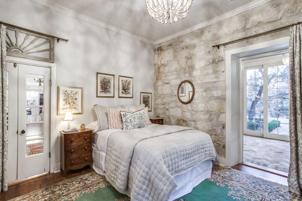 A cozy bedroom with a stone accent wall, white bedding, wooden nightstands, framed artwork, and a round mirror styled by an interior decorator Boerne. Double doors open to a sunlit patio while a green patterned rug covers the wooden floor.