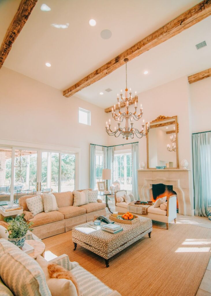 Bright, airy living room with beige sofas, patterned ottoman, large windows, and exposed wooden ceiling beams. Designed by a top interior design company in Boerne TX, this space features a chandelier and fireplace with a mirror above.
