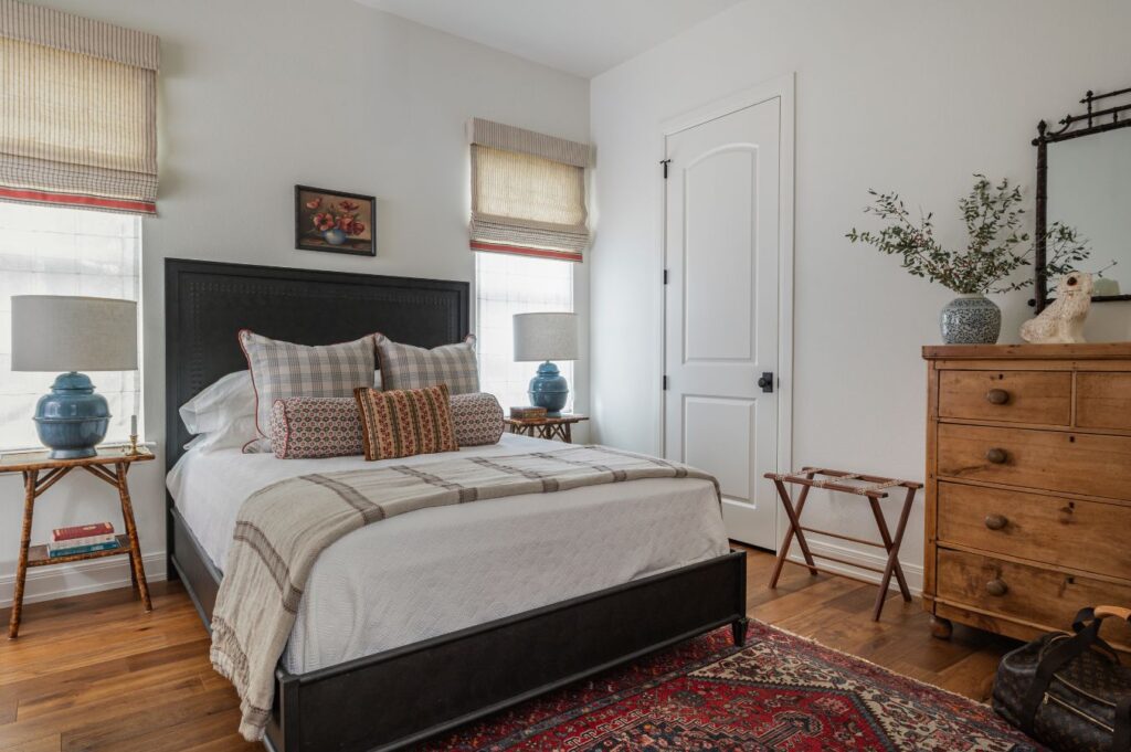 A cozy bedroom with a black-framed bed, plaid pillows, two blue lamps on small side tables, a wooden dresser with a mirror, a patterned rug, and woven blinds on the windows letting in natural light.