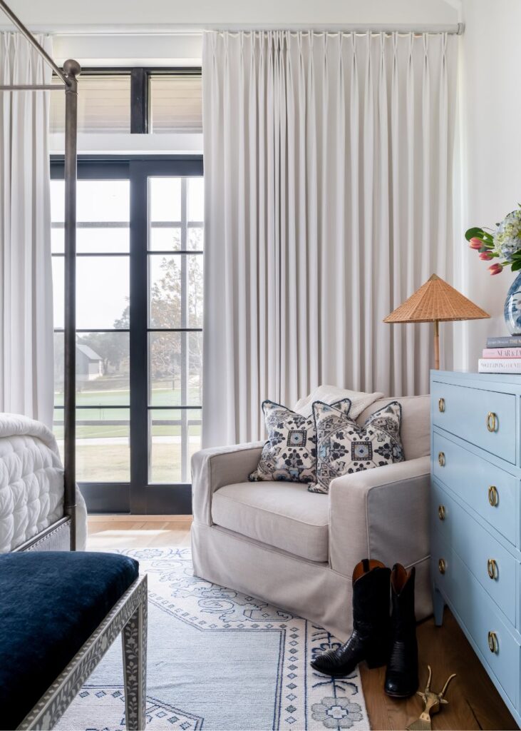 A cozy bedroom corner styled by an interior decorator Boerne, with a beige armchair holding two patterned pillows, a blue dresser with books and flowers, light curtains, a window view, black boots on a blue rug, and part of a bed visible.
