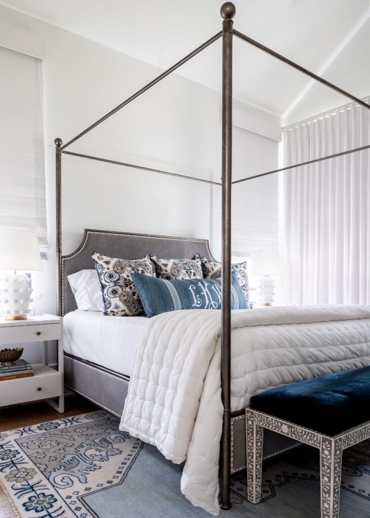 A modern bedroom by an interior designer Boerne TX features a metal four-poster bed, crisp white bedding, blue and white pillows, a velvet bench, sleek nightstands with lamps, and large windows dressed in airy curtains.