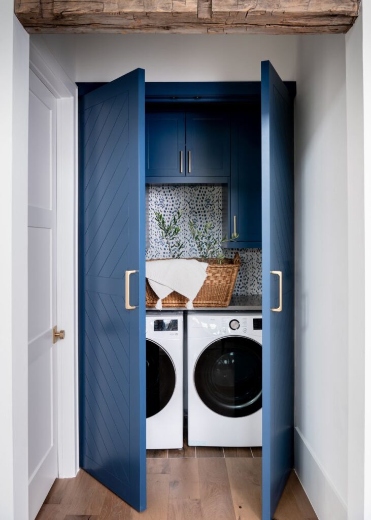 A compact laundry nook with blue double doors, designed by an interior decorator Boerne, features a front-loading washer and dryer, blue cabinetry above, a blue and white backsplash, wicker baskets, and potted plants for decor.