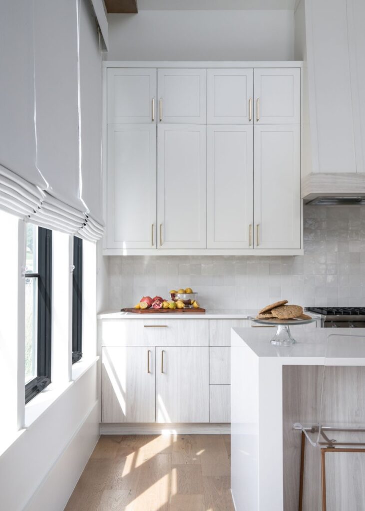 Bright modern kitchen with white cabinets, light wood floors, and large windows with white blinds. Styled by an interior designer Boerne TX, the countertop holds fruits and bread for a clean, airy feel.